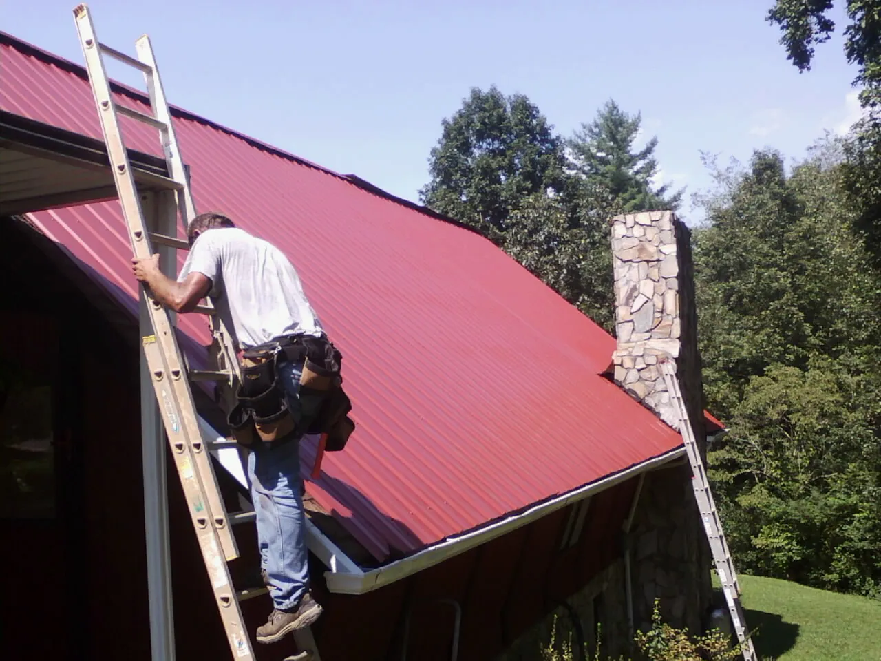 Roofer on a ladder beside a red metal roof and stone chimney — Soffit & Fascia Repair by Roofing Brothers in Almo